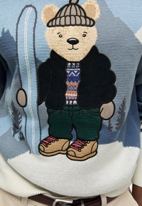 Sweter wydziergany z grafiką misia w zimowej scenerii, noszącego ciemną kurtkę puchową, wzorzysty sweter, zielone spodnie i brązowe buty.