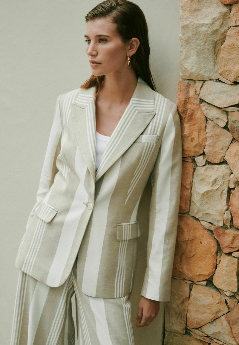Jeune femme aux cheveux mouillés portant un blazer à rayures verticales beige et blanches et un pantalon large assorti, debout contre un mur en pierre.