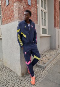 Marineblå træningsdragt med gule, pink og blå detaljer; inkluderer en jakke med lynlås og matchende bukser. Skarpe orange sneakers. Urban miljø.