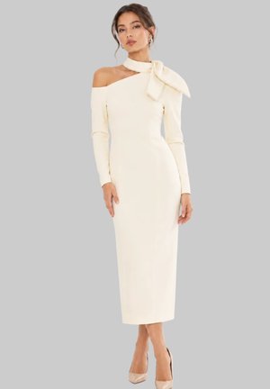 Frau in einem figurbetonten cremefarbenen Midi-Kleid mit schulterfreiem Design, langen Ärmeln und großem Halsband, die vor einem einfarbigen grauen Hintergrund posiert.