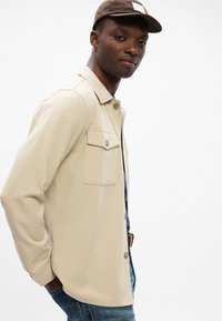 Μπεζ πουκάμισο με κουμπιά και δύο τσέπες στο στήθος, καθώς και γιακά. Το ύφασμα είναι λείο και ελαφρύ. Φοριέται με μπλε τζιν και καφέ καπέλο.