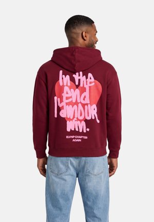 Un hombre luce una sudadera con capucha de color burdeos, con un corazón rosa y letras en la espalda, combinada con unos vaqueros azul claro.