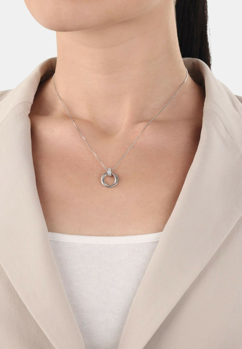 Silverhalsband med sammanflätade cirkulära länkar, med en strukturerad accentdetalj, bärs över en enkel vit topp under en beige kavaj.