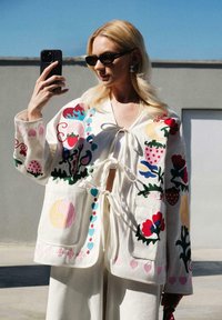 Valge tikandiga jakk, millel on värvikad lille- ja puuviljamustrid, lipsuga kinnitamine ja kaks esist pocketit. Sile tekstuur, mugav lõige.