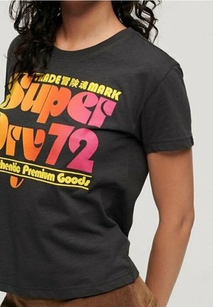 Kvinde iført en sort Superdry 72 T-shirt med pink, orange og gul tekst på fronten mod en grå baggrund.