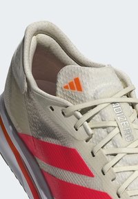 Světlé béžové sportovní boty s texturovaným svrškem, obsahují růžové a oranžové akcenty a bílé šňůrky. Výrazné logo Adidas na jazyku.