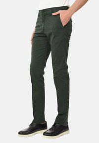 Pantaloni di cotone verdi con vestibilità slim, tasche frontali e chiusura con bottone. Abbinati a scarpe nere con suola bianca.