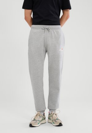 Persoon die lichtgrijze joggingbroek met aantrekkoord, zwart shirt en beige sneakers met groene en rode accenten draagt, staand tegen een effen achtergrond.
