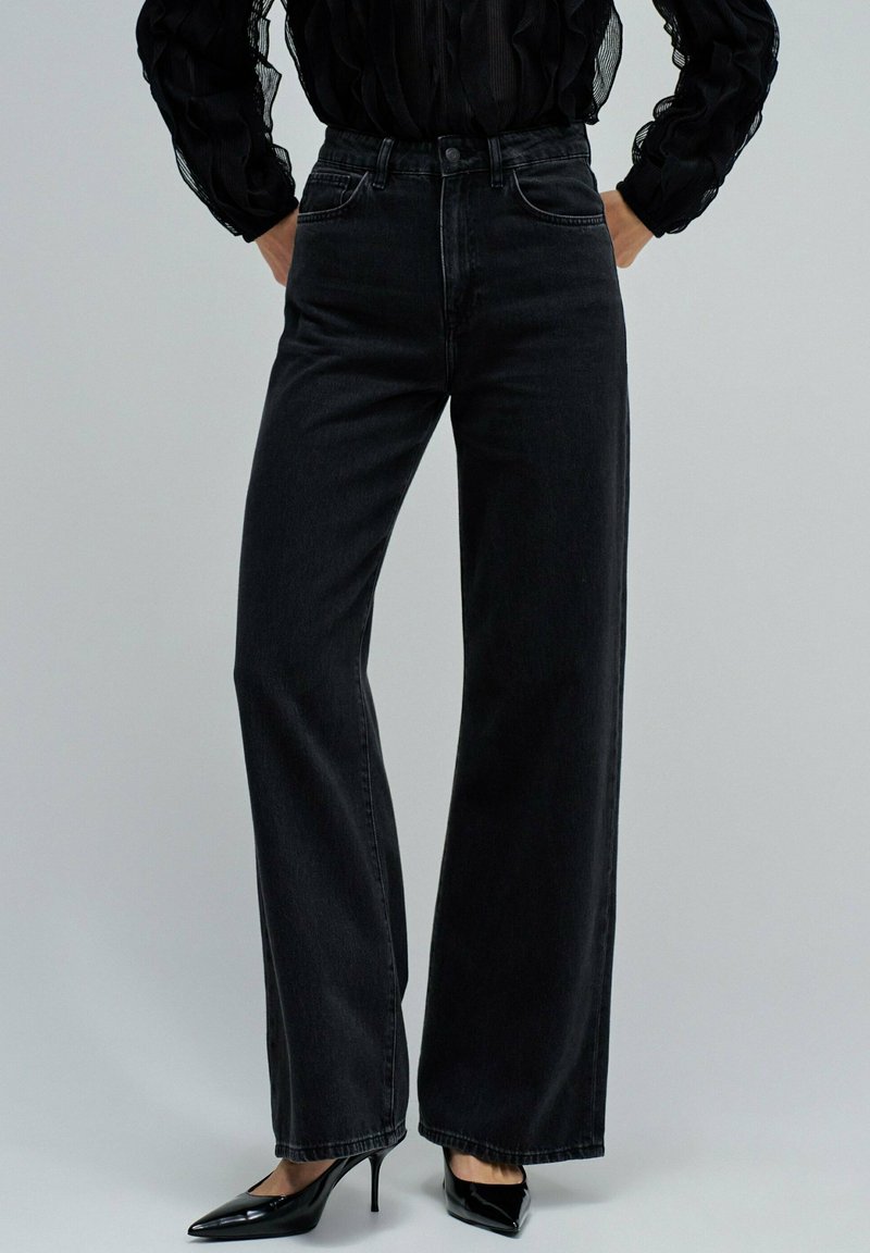 Jeans de denim de pernera ancha de talle alto en negro, con un diseño clásico de cinco bolsillos y costuras sutiles. Combinados con tacones negros de punta.