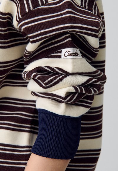 Maglione a righe con linee crema e marroni, dotato di un polsino a costine blu navy e una piccola etichetta ricamata bianca con la scritta "Claudie". Texture morbida.