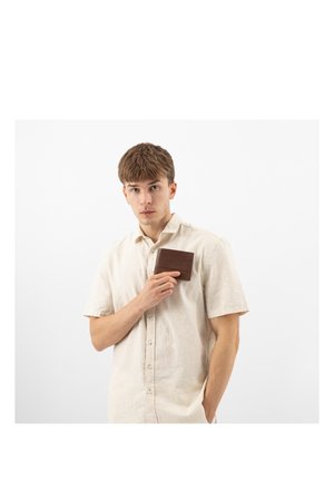 Ein junger Mann trägt ein beiges Hemd und hält eine braune Lederbrieftasche nahe an seiner Brust, vor einem schlichten weißen Hintergrund stehend.