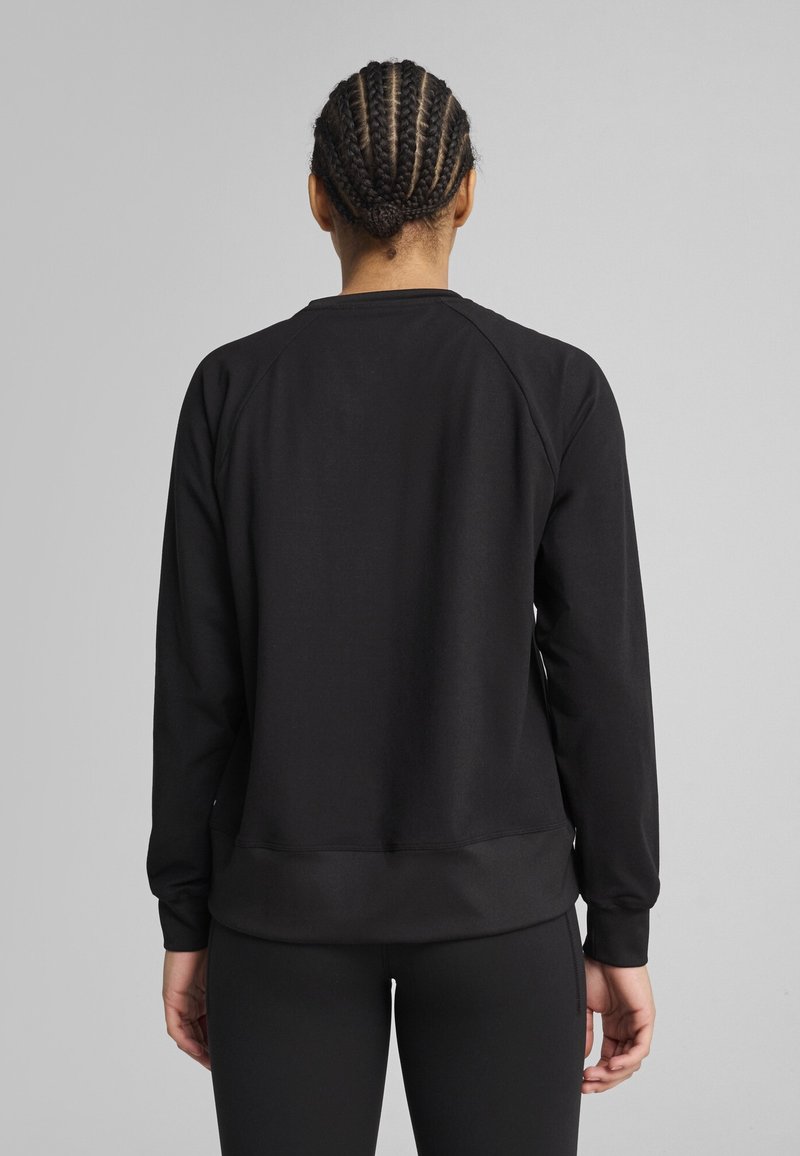 Personne aux cheveux tressés portant un haut noir uni à manches longues et un pantalon noir, debout dos à un fond gris.