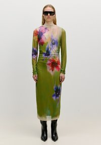 Langärmeliges Kleid mit grünem Grundton, das ein buntes Blumenmuster in Lila, Rot und Gelb zeigt. Glattes Material, figurbetonte Passform. Schwarze Stiefel.