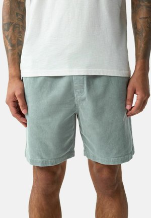 Man wearing light green corduroy shorts and a white t-shirt, showing tattooed arms and hands resting by sides.