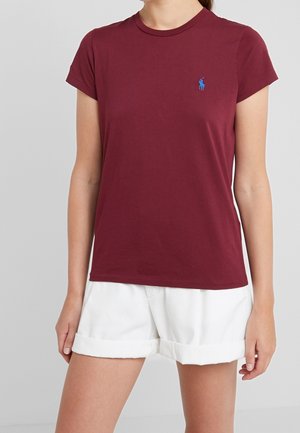 Bordeauxrotes Baumwoll-T-Shirt mit kurzen Ärmeln und Rundhalsausschnitt. Verfügt über ein kleines, blau gesticktes Logo auf der Brust. Wird mit weißen hochgekrempelten Shorts getragen.