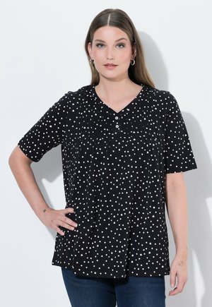 Femme aux cheveux bruns lisses porte un chemisier noir à manches courtes avec des motifs de cœurs blancs et un jean bleu, debout avec une main sur la hanche.