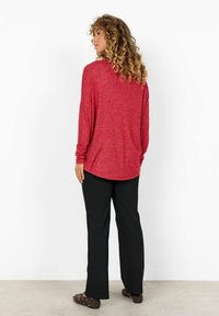 Langsleeve, rood gemêleerd top met een gebogen zoom, gecombineerd met zwarte broeken. Het model draagt platte schoenen met studded accenten.