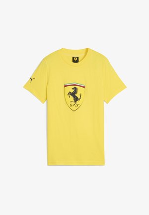 Kollane t-särk, mille keskel on Ferrari logo, must hüplev hobune ja värvilised triibud embleemi kohal.