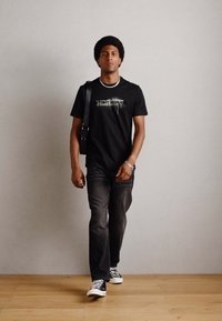 Tricou negru cu logo, blugi închisi și adidași alb-negru; modelul ține o geantă crossbody neagră cu un umăr. Fundal din podea din lemn.