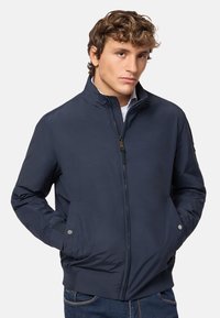 Giacca bomber blu navy con collo alto, zip frontale, due tasche laterali, hardware minimale e polsini a costine. Tessuto liscio e leggero.
