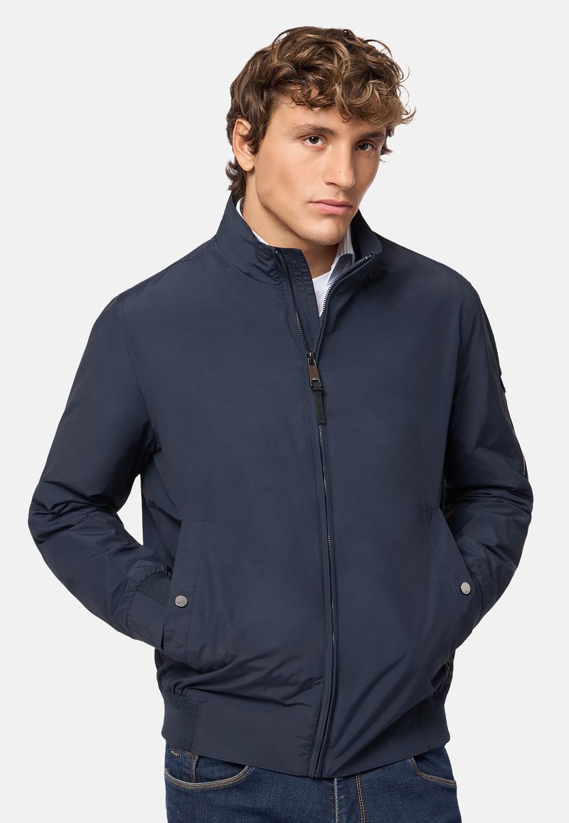 Giacca bomber blu navy con collo alto, zip frontale, due tasche laterali, hardware minimale e polsini a costine. Tessuto liscio e leggero.