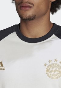Dres FC Bayern München s bílým tělem a tmavě námořnickými rukávy typu raglan. Obsahuje zlaté brandingy a vyšívané logo se hvězdnými akcenty.