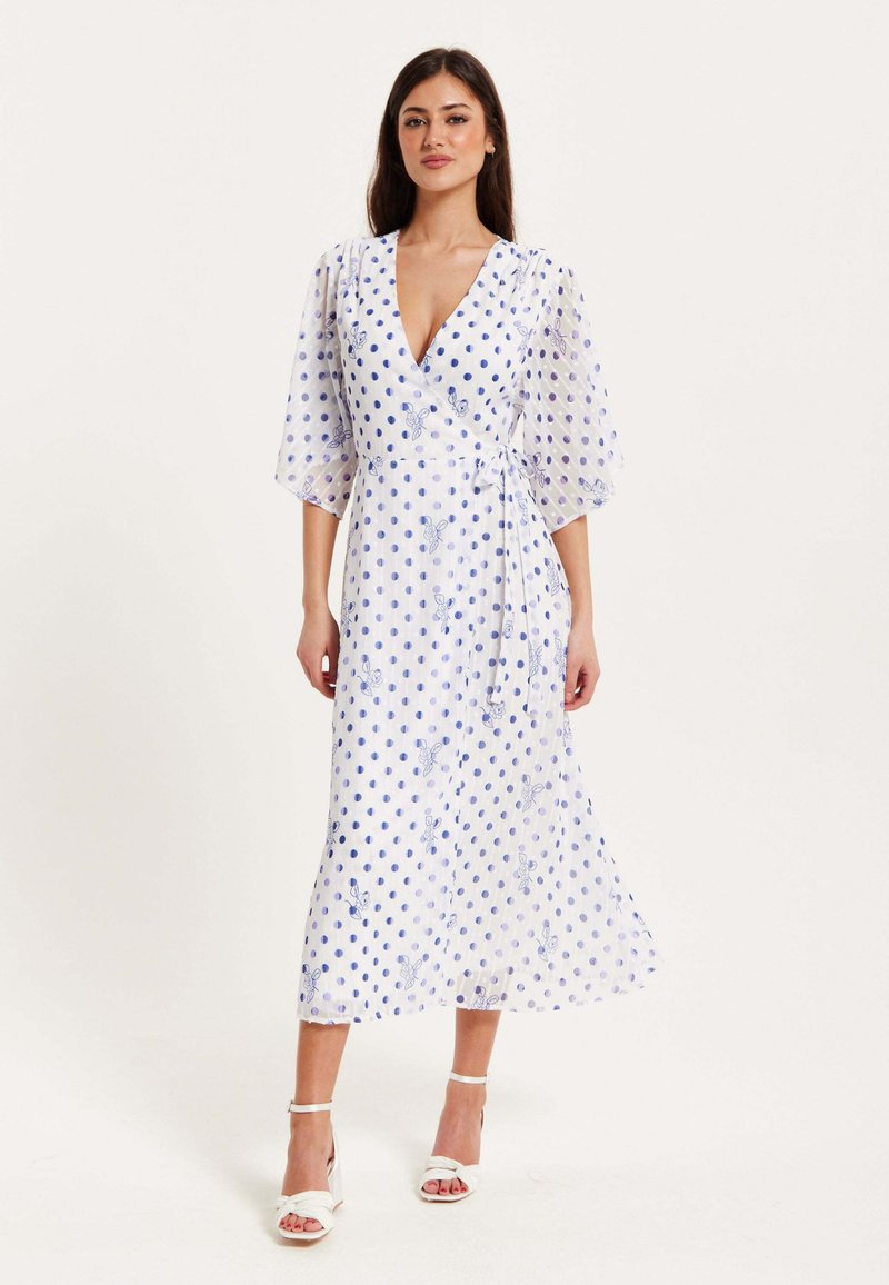 White wrap dress featuring blue polka dots and butterfly embroidery. Flowy fabric, deep V-neckline, and three-quarter puff sleeves.