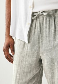 Pantaloni din in gri deschis, cu talie elastică și șnur. Material texturat și design cu picioare largi. Camasa albă cu nasturi, parțial vizibilă.