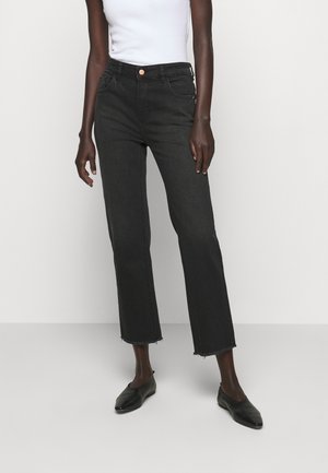 Model draagt zwarte cropped jeans met hoge taille en onafgewerkte zoom, gecombineerd met een wit mouwloos topje en zwarte puntige platte schoenen.