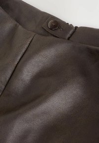 Indumento in pelle marrone scuro con un linguetta in vita dotata di bottoni e dettagli di cucitura visibili vicino al margine superiore.