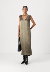 Robe en satin vert olive avec décolleté en V, sans manches et coupe droite. Portée avec des bottes noires à bout pointu et un petit sac à main noir.