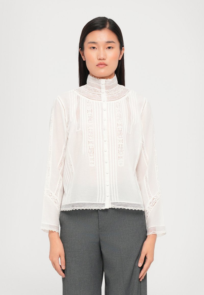 Blusa blanca transparente con intrincados detalles de encaje, rayas verticales y un cuello alto, con cierres de botones en la parte delantera.