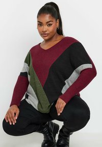 Maglione oversize a righe rosso, nero, verde e grigio con scollatura a V e maniche lunghe. Indossato con pantaloni neri e stivali.