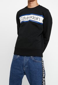 Czarna bluza z biało-niebieskim paskiem z napisem "Calvin Klein". Posiada okrągły dekolt i ściągacze przy rękawach, zestawiona z niebieskimi dżinsami.