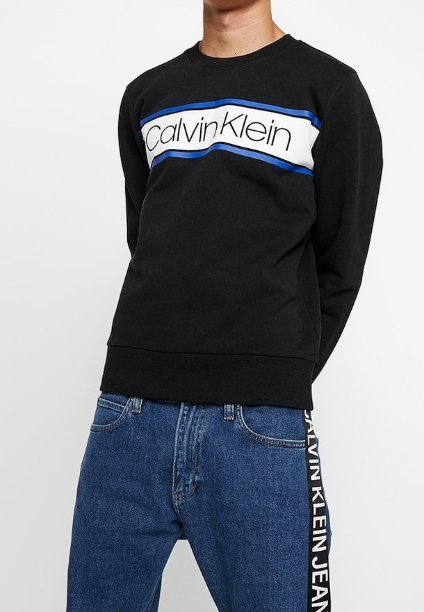 Czarna bluza z biało-niebieskim paskiem z napisem "Calvin Klein". Posiada okrągły dekolt i ściągacze przy rękawach, zestawiona z niebieskimi dżinsami.