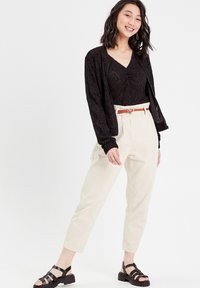 Cardigan en dentelle noire porté sur un top noir à col en V, associé à un pantalon crème et une ceinture marron. Des sandales noires à brides complètent l'ensemble.