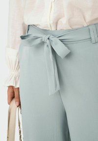 Pantalon taille haute bleu clair avec une finition texturée, doté d'une ceinture nouée en forme de nœud à la taille. Un chemisier de couleur crème est partiellement visible.