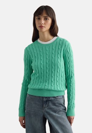 Young woman wearing a mint green cable-knit sweater over a white shirt and blue jeans, standing against a plain background.