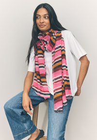 Sciarpa multicolore con strisce a zigzag rosa, marrone e nere, realizzata in tessuto leggero, elegantemente drappeggiata intorno al collo di una modella.