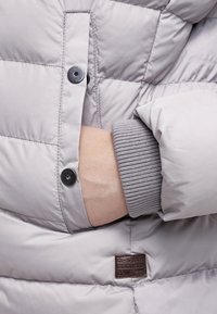 Giacca piumino grigio chiaro con texture trapuntata, dotata di bottoni neri e polsini a costine. Include una toppa con logo marrone sul lato.