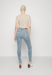 Jeans ajustados en denim claro, con una cintura alta, dos bolsillos traseros y una etiqueta de cuero marrón en la parte trasera. Combinados con una blusa beige y tacones.