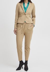 Beige kavaj med en figurnära passform, tillsammans med matchande smalbyxor och en teal knapp-up skjorta. Svarta spetsiga klackar fullbordar outfiten.