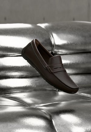 Dunkelbrauner Lederloafer ruht auf einem glänzenden silbernen, getufteten Kissen.