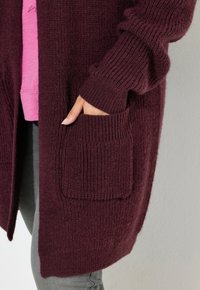 Cardigan lavorato a maglia color bordeaux con texture a coste, design aperto davanti e due tasche frontali; indossato sopra un top rosa con un fondo grigio.