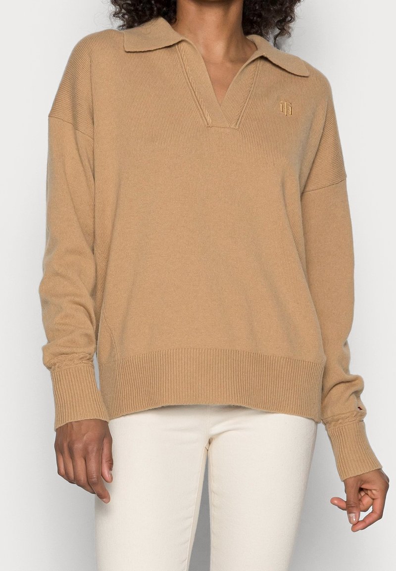 Pull-over en tricot beige avec un col à revers, des poignets et un ourlet côtelés. Dispose d'un petit logo sur la poitrine et d'une coupe décontractée.