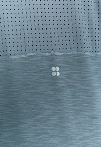 Helles blaues Sportoberteil mit einer perforierten Textur oben und glattem Stoff darunter, ausgestattet mit einem dezenten Logo in der unteren rechten Ecke.