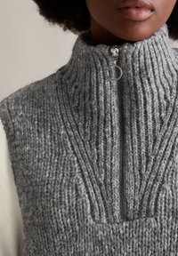 Nahaufnahme einer Person, die einen grauen, dick gestrickten ärmellosen Pullover mit einem silbernen Reißverschluss und einem runden Zuggriff trägt.