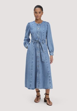 Femme portant une robe en jean à manches longues avec des boutons sur le devant et une ceinture à la taille, associée à des sandales à talons carrés marron à brides.