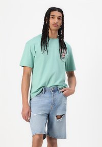Tricou cu mânecă scurtă de culoare verde mentă cu imprimeu grafic pe piept, asociat cu pantaloni scurți din denim albastru deschis cu detalii distruse și o ruptură mare.