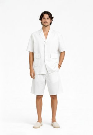 Uomo con capelli scuri e ricci che indossa un blazer bianco a maniche corte, pantaloncini coordinati fino al ginocchio e scarpe slip-on su sfondo bianco.